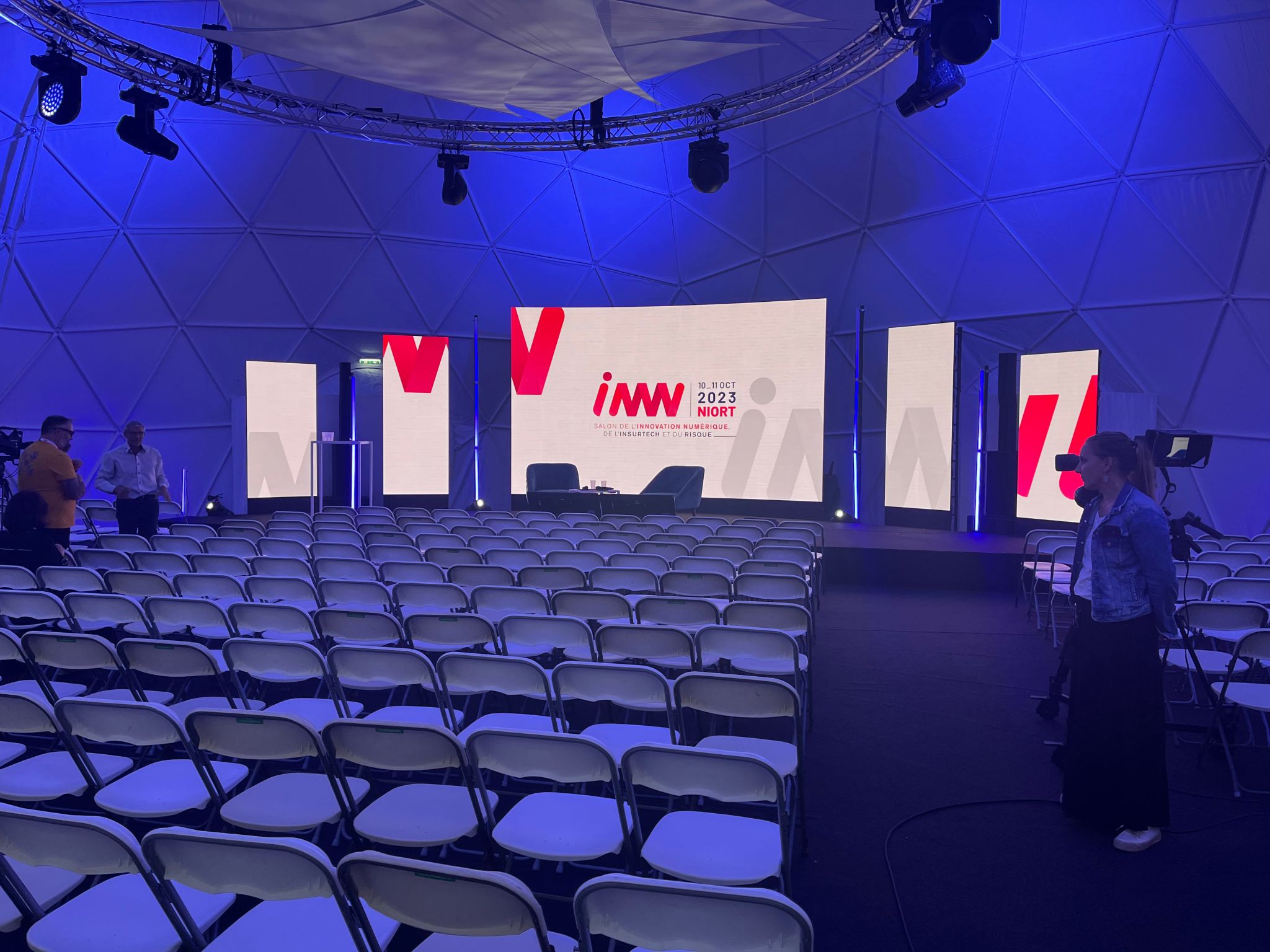 Une salle vide préparée pour l'événement INNN 2023 à Niort, avec des écrans LED et une scène installés, éclairés par des lumières bleues. Prestation technique assurée par TEDELEC Event.